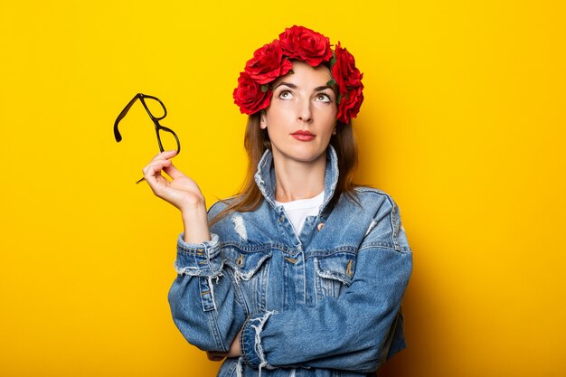 Jonge vrouw in een spijkerjasje en een krans van rode bloemen op haar hoofd houdt een bril op een gele muur.