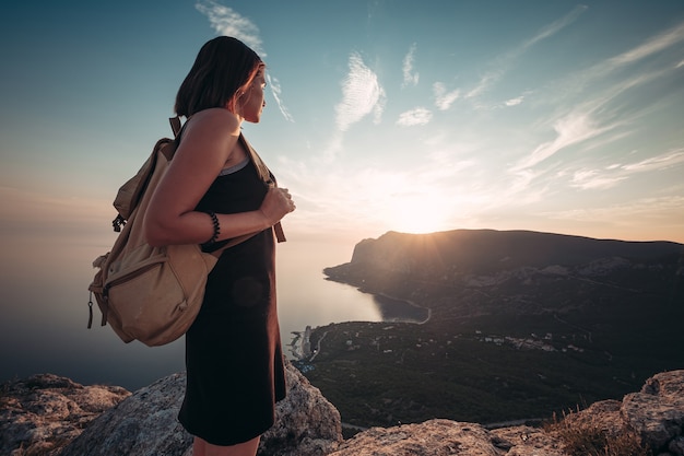 Jonge vrouw in een sportjurk met rugzak geniet van het uitzicht op de bergen en de zee bij zonsondergang. Reizen en actieve levensstijl concept. glamping