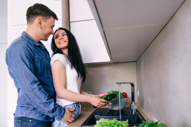 Jonge vrouw in een T-shirt wast groenten in de keuken De man omhelst het meisje zachtjes