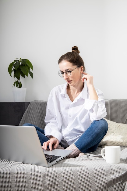 Jonge vrouw in een wit overhemd met een laptop aan het werk terwijl ze op de bank zit