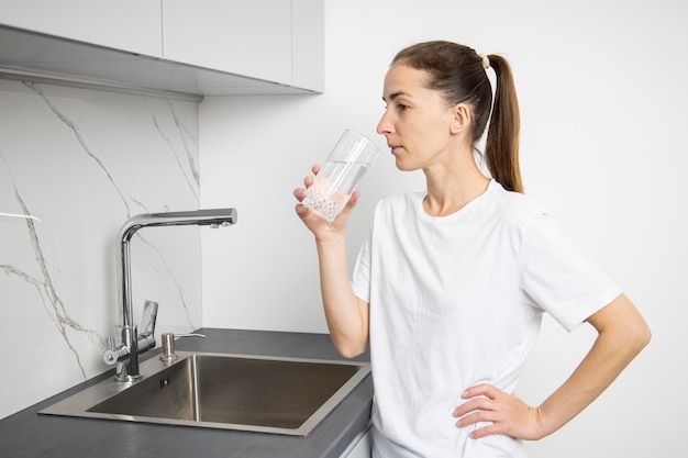 Jonge vrouw in een wit t-shirt in de keuken drinkt water uit een glazen beker