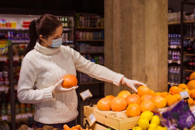 Jonge vrouw in een witte gebreide trui met een beschermend masker die appels kiest in een plaatselijke winkel Voedsel kopen in een plaatselijke winkel om kleine bedrijven te ondersteunen