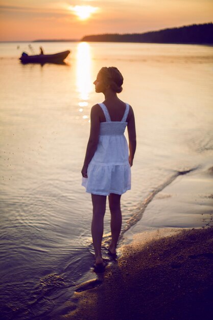 jonge vrouw in een witte jurk die bij de zee staat bij zonsondergang