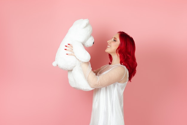 Jonge vrouw in een witte jurk en met rood haar die naar een grote witte teddybeer kijkt