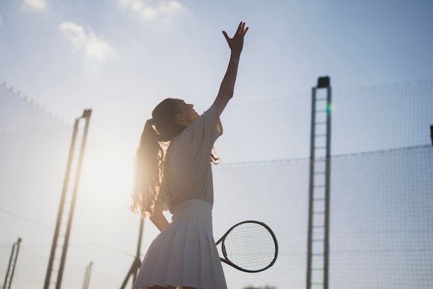jonge vrouw in een witte tennis outfit staat op een tennisbaan klaar om de bal te serveren de zon schijnt helder achter haar het creëren van een prachtig silhouet