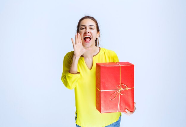 Jonge vrouw in geel shirt die iemand belt of uitnodigt om een rode geschenkdoos te presenteren