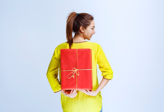 Jonge vrouw in geel shirt verbergt een rode geschenkdoos achter zichzelf