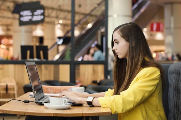 Jonge vrouw in gele jas aan het werk met de computer in het café freelance werk