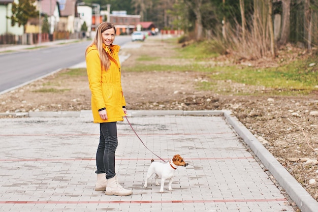 Jonge vrouw in gele jas, glimlachend en kijkend naar de camera, met Jack Russell Terriër hond aan een roze leiband, wazig huizen op de achtergrond