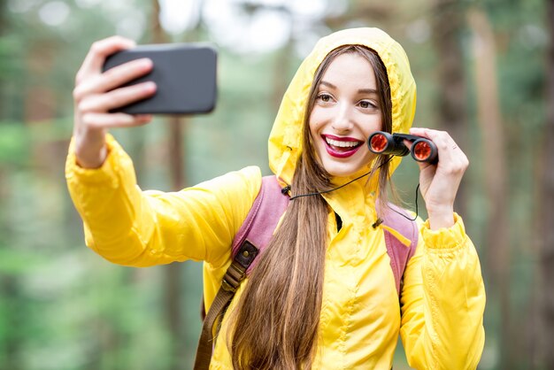 Jonge vrouw in gele regenjas die selfie portret neemt tijdens het wandelen met een verrekijker en rugzak in het groene bos