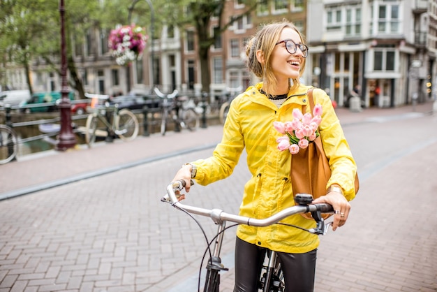 Jonge vrouw in gele regenjas met tas en bloemen fietsen in de stad Amsterdam