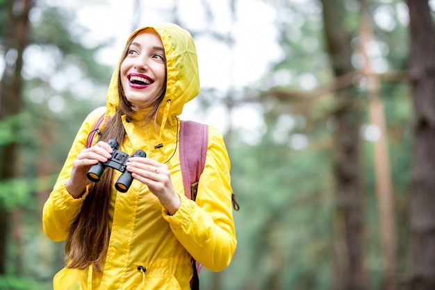 Jonge vrouw in gele regenjas op zoek met een verrekijker in het groene dennenbos. Vogels kijken in het bos