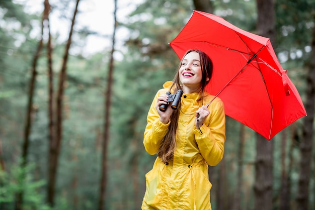 Jonge vrouw in gele regenjas wandelen met verrekijkers en rode paraplu in het dennenbos