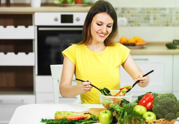Jonge vrouw in gele t-shirt die groentesalade roert in een transparante kom die binnen is geschoten in moderne witte keuken