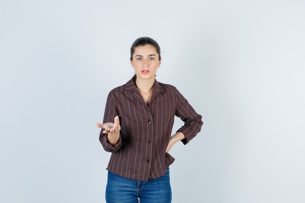 Jonge vrouw in gestreept shirt, jeans met hand op heup, hand op vragende manier uitrekkend en verbaasd kijkend, vooraanzicht.