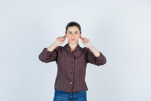 Jonge vrouw in gestreept shirt, jeans met handen achter de oren en er serieus uitziend, vooraanzicht.