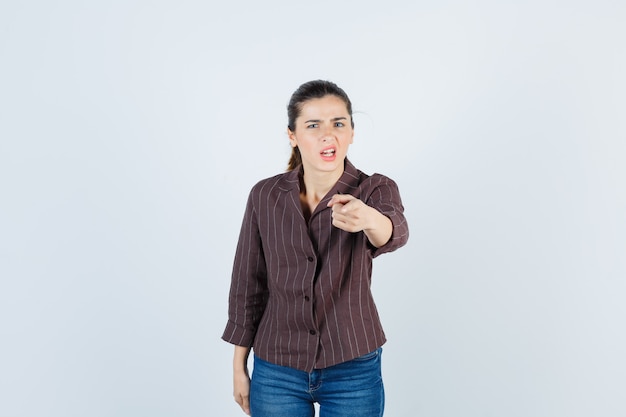 Jonge vrouw in gestreept shirt, jeans wijzend naar voren met wijsvinger, grimassen en kijken gehaast, vooraanzicht.