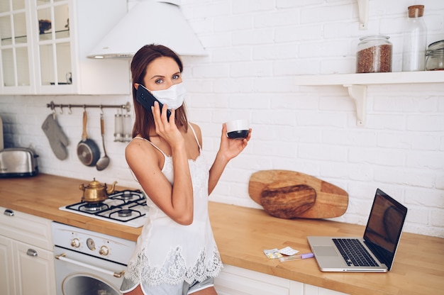 Jonge vrouw in gezichtsmasker telefoon spreken en werken op een laptop met koffie in de ochtend. Thuisquarantaine tijdens Covid-19 pandemisch Corona-virus.