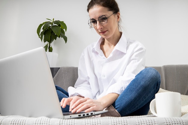 Jonge vrouw in glazen met laptop met koffie zittend op de bank