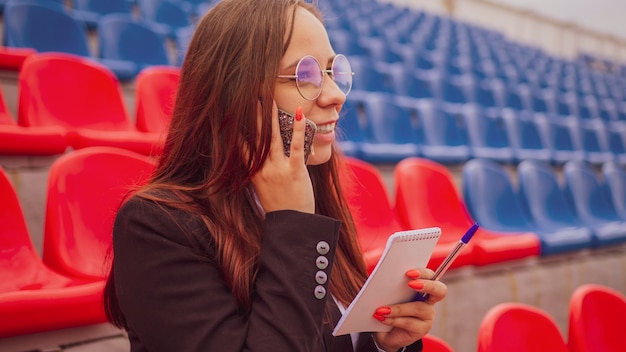 Jonge vrouw in glazen met notitieblokpen praten op mobiele telefoon zittend op stadion tribunes