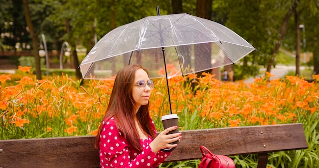 Jonge vrouw in glazen met transparante paraplu koffie drinken uit papieren beker zittend op bankje bij regenachtig weer Ontspannen vrouw in rode jurk opwarming van de aarde met warme drank op wandeling rusten in stadspark