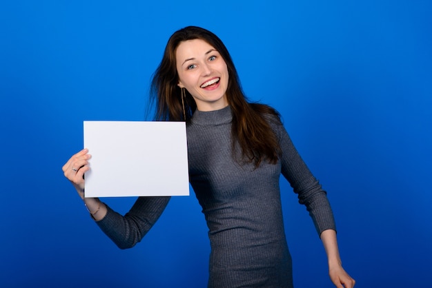 Jonge vrouw in grijs shirt met een vel papier en kijken naar de camera, glimlachend. blauwe achtergrond geïsoleerd
