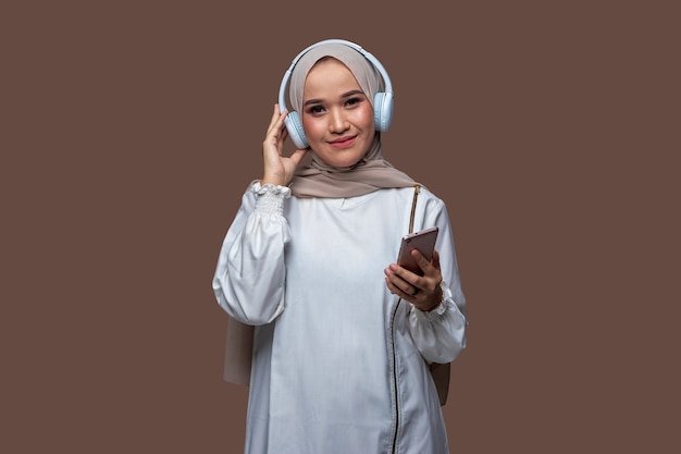 Jonge vrouw in hijab die zich voordeed met een koptelefoon op en een mobiele telefoon vasthoudt