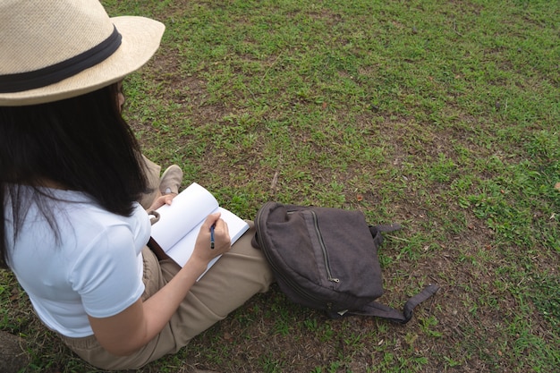Jonge vrouw in hoed die op notitieboekje met potlood op gras in openluchtpark schrijft.