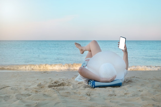 Jonge vrouw in hoed met mobiele telefoon op het strand. freelance meisje liegt