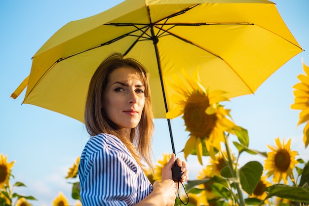 Jonge vrouw in jurk verstopt voor de zon onder een gele paraplu op een zonnebloem veld.