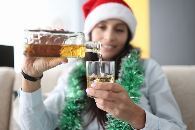 Jonge vrouw in kerstman hoed en klatergoud om haar nek giet alcohol uit fles in glas met ijs