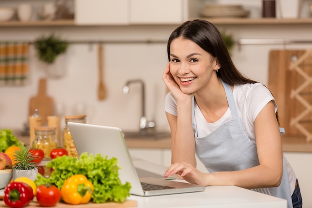 Jonge vrouw in keuken met laptop zoekt recepten, glimlachend. Voedsel blogger concept