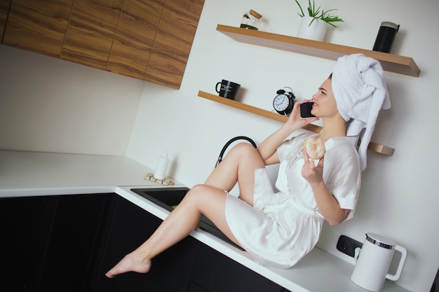 Foto jonge vrouw in keuken tijdens quarantaine. aantrekkelijk meisje zitten en praten over de telefoon na het douchen houd donut in de hand. sexy meisje brengt tijd door in de keuken.