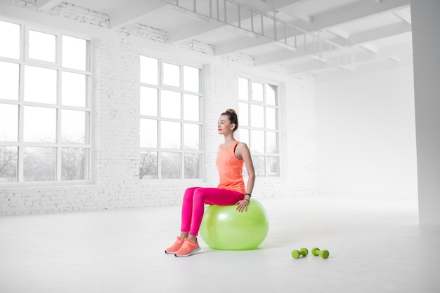 Jonge vrouw in kleurrijke sportkleding die fitness doet met fitnessbal in de witte sportschool