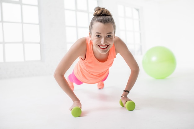 Jonge vrouw in kleurrijke sportkleding die push-ups maakt met halters en fitnessbal op de achtergrond