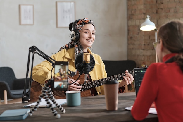 Jonge vrouw in koptelefoon die gitaar speelt en een lied zingt tijdens de lucht in de radiostudio met radio-dj die tegenover haar zit