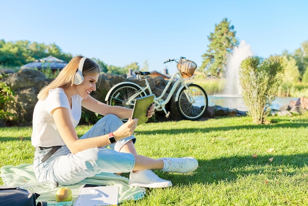 Jonge vrouw in koptelefoon met digitale tablet op zoek in het scherm van gadget, video, film, oproep. Vrouw zittend op groen gazon in park op zonnige zomerdag