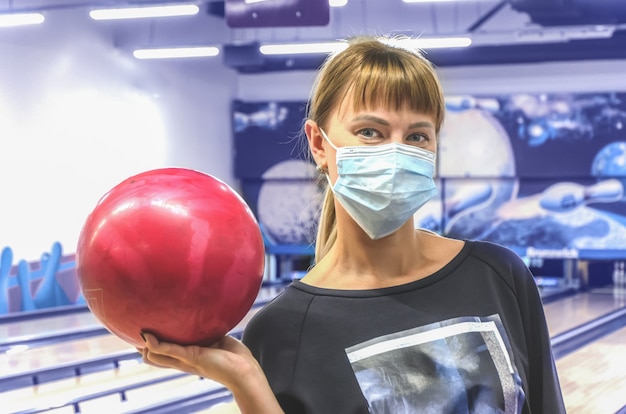 Jonge vrouw in medisch masker in de holdingskegelenbal van de bowlingclub van sporen en spelden. Actieve vrijetijdsbesteding. Sportieve gezinsactiviteiten. Wazig getinte afbeelding met selectieve aandacht