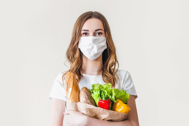 Jonge vrouw in medische masker heeft een eco-papieren zak met voedsel, fruit en groenten, peper, stokbrood, sla, veilige online slimme levering, coronovirus, quarantaine, pandemie, blijf thuis concept