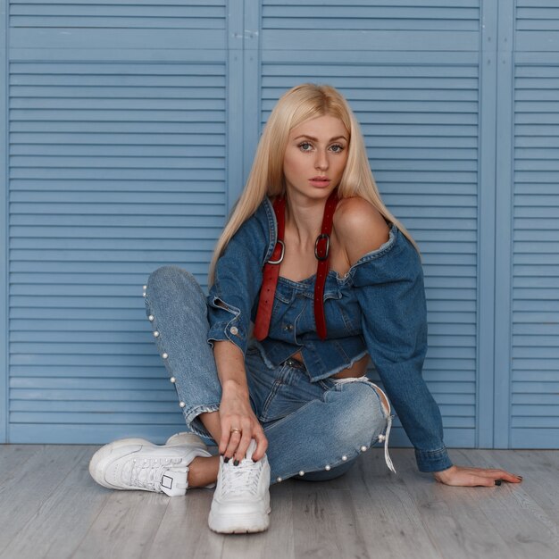 Jonge vrouw in mode denim jurk met riemen en witte sneakers zitten in de buurt van een blauwe houten muur