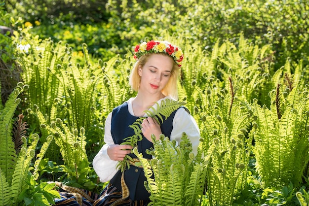 Jonge vrouw in nationale kleding en krans tegen een groene varen Ligo Letse vakantie