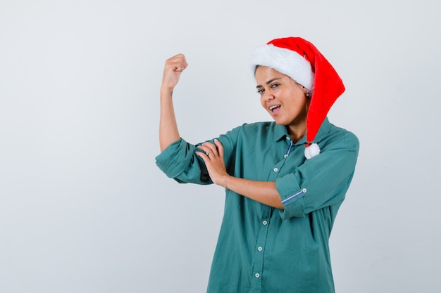 Jonge vrouw in overhemd, kerstmuts die spieren van de arm toont en er zelfverzekerd uitziet, vooraanzicht.