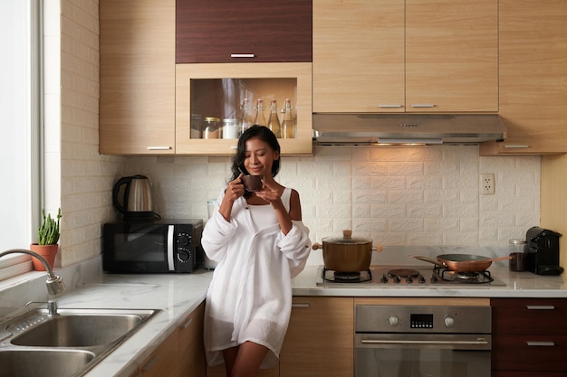 Jonge vrouw in pyjama die aan het aanrecht staat en koffie drinkt en geniet van de nieuwe ochtend