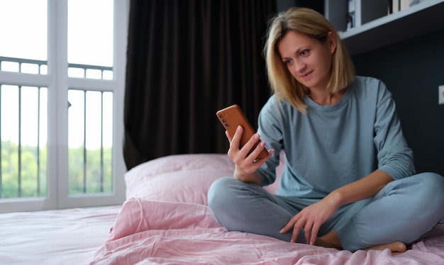 Jonge vrouw in pyjama zit in bed met mobiele telefoon in de hand