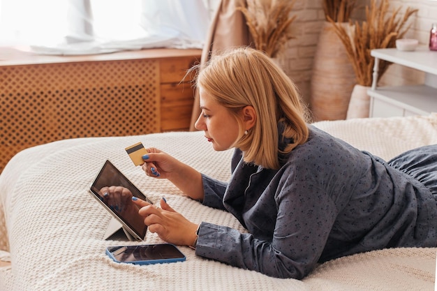 Jonge vrouw in pyjama zit op het bed met een computer en een telefoon doet een online betaling