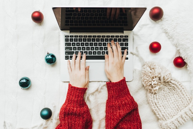 Jonge vrouw in rode gebreide trui typen op laptop op het witte bed met witte deken versierd met rode en blauwe kerstballen