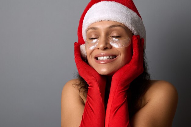 Jonge vrouw in Santa carnaval outfit, met glanzende gladmakende vlekken onder haar ogen, houdt haar handen bij haar gezicht, glimlacht met een mooie brede glimlach en poseert met gesloten ogen op een grijze achtergrond