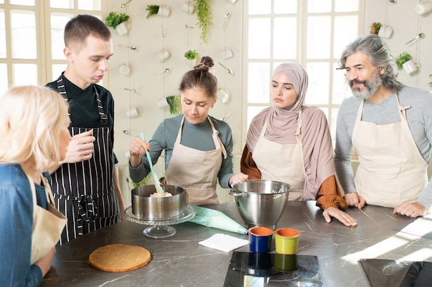 Jonge vrouw in schort die zelfgemaakte botercrème op zelfgemaakte cake of ander dessert in een grote metalen kom verspreidt tijdens het koken