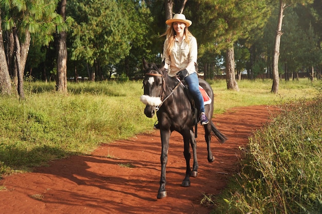 Jonge vrouw in shirt en strohoed, klein zwart paard berijdend in het park, wazige achtergrond met huizen en bomen