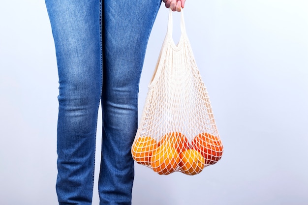 Jonge vrouw in spijkerbroek is string tas met verschillende citrus op grijze achtergrond. Concept zero waste, milieuvriendelijk.